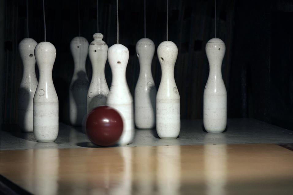 Eine rotbraune Bowlingkugel rollt auf eine Reihe weißer Kegel auf einer Bowlingbahn zu.