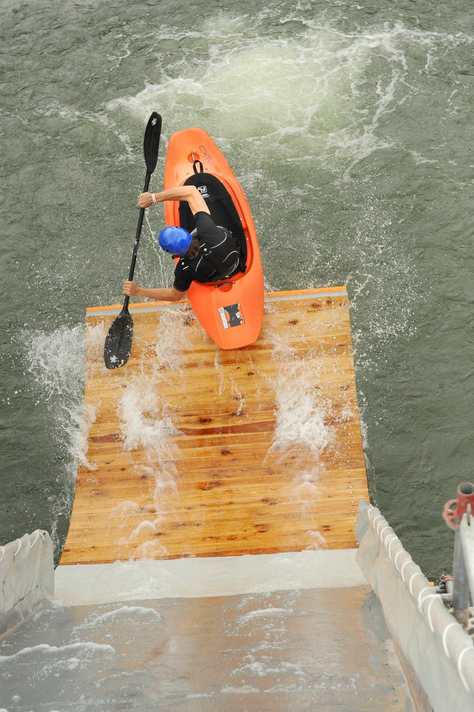Paddler in orangenem Kajak steuert über eine Holzrampe ins Wasser, spritzendes Wasser um ihn herum.