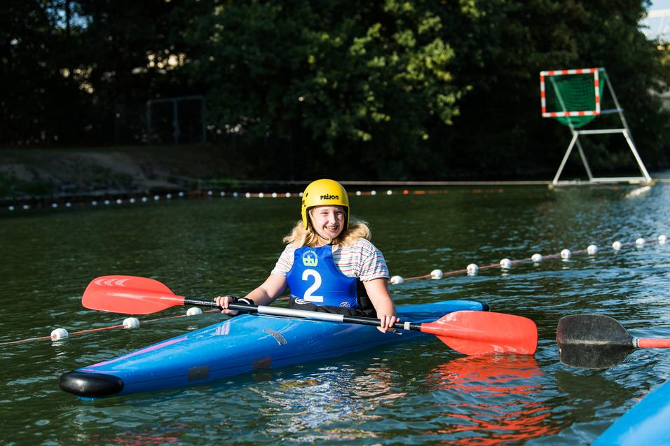 Junge Frau im blauen Kajak mit rotem Paddel lächelt auf einem Wasserweg, im Hintergrund ein Tor fürs Wasserballspiel.